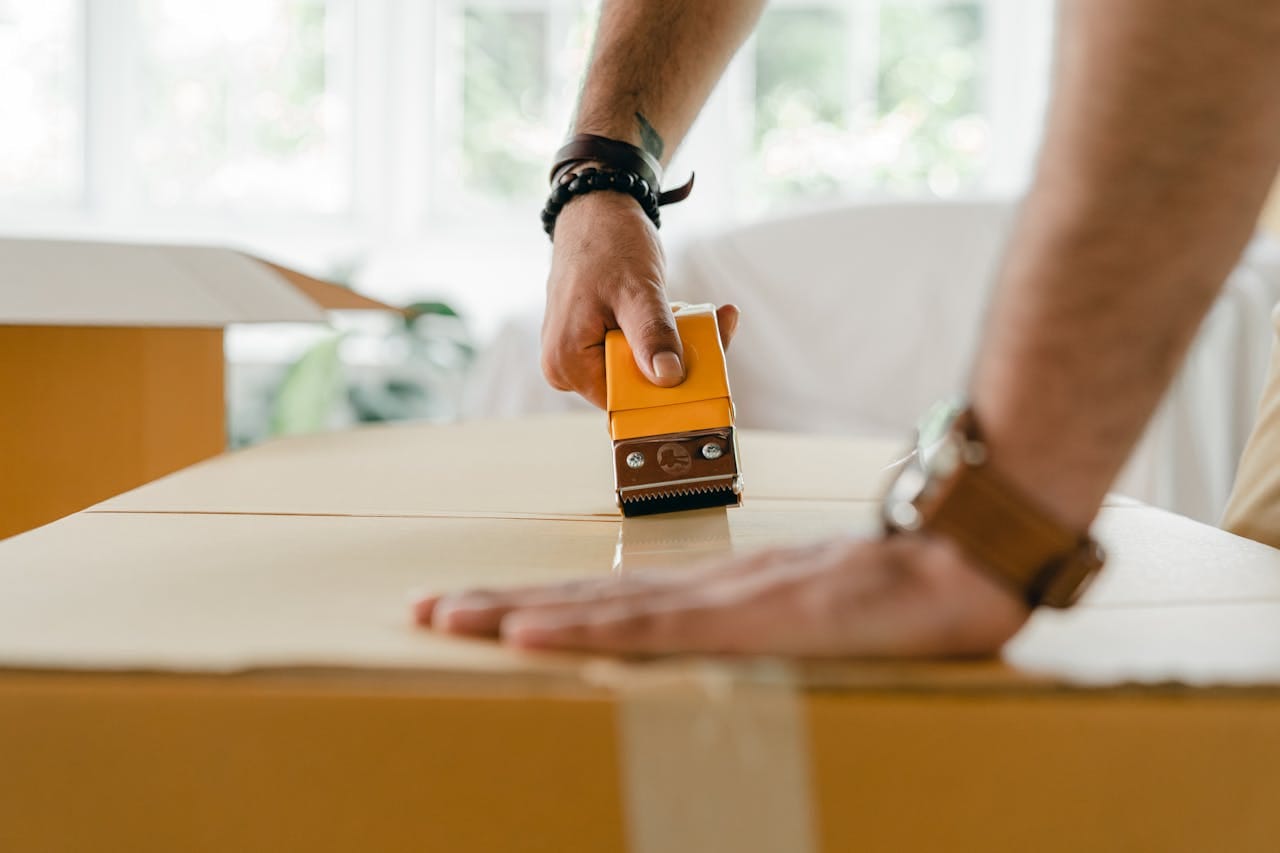 Side view of crop anonymous man using scotch tape dispenser for packing big carton box for moving personal items to new apartment